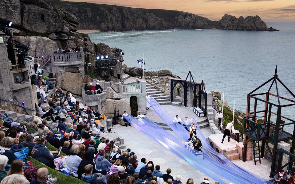 Actors performing on the Minack stage with audience watching against an evening sky