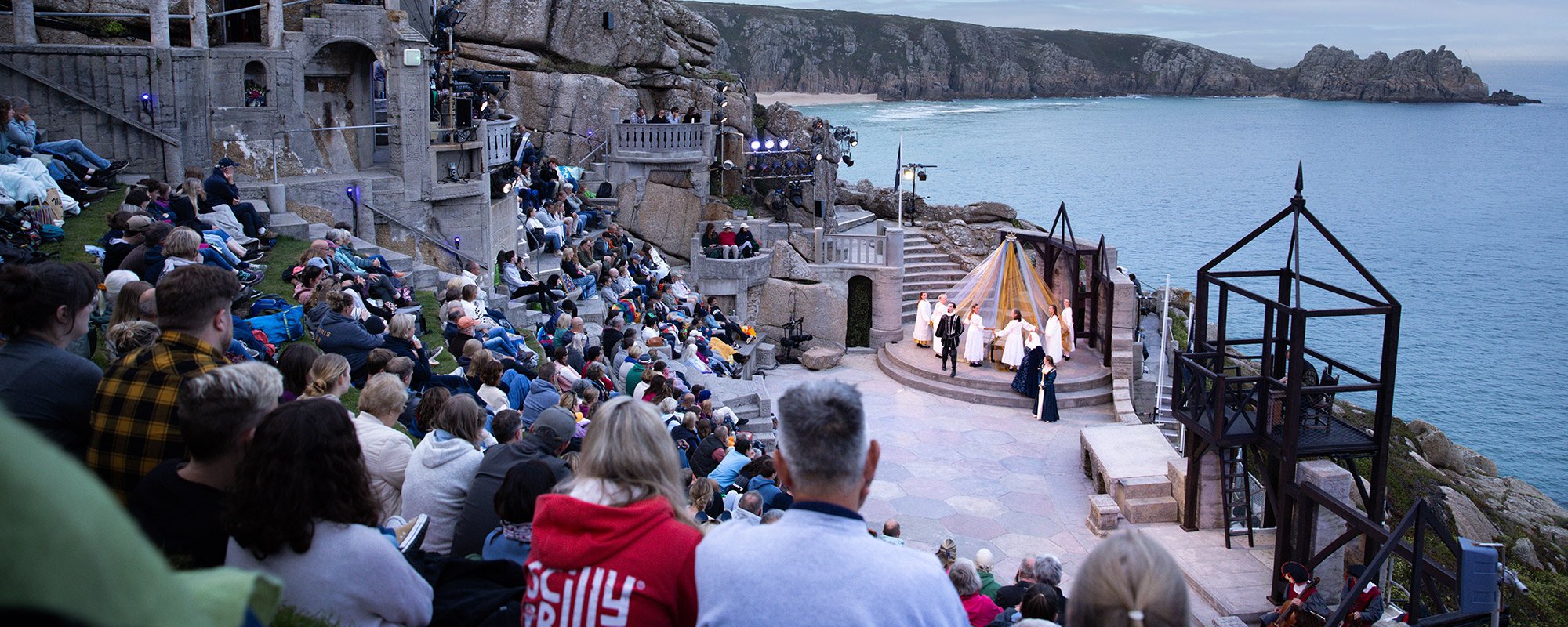 Actors performing on the Minack stage with audience watching in the early evening